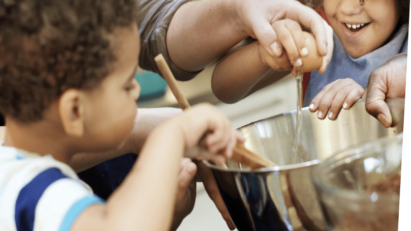 children with curly hair and light brown skin gleefully helping an adult add ingredients to a bowl for mixing