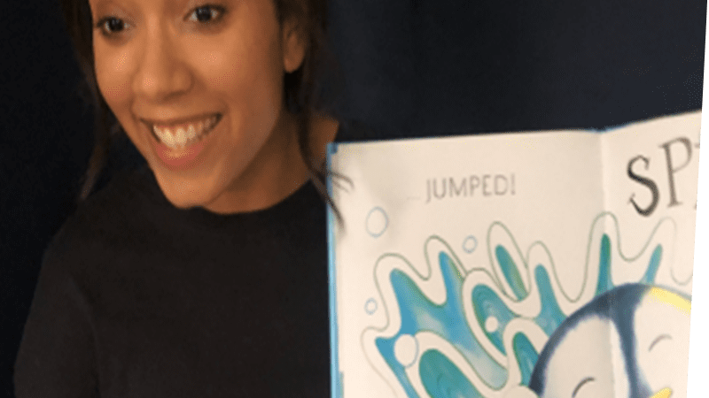 A woman smiling and displaying a story book. The open book shows an illustrated penguin jumping into water with a splash.