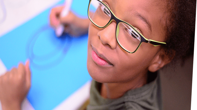 Child looking up at the camera from her work drawing on a blue sheet of paper with a red marker