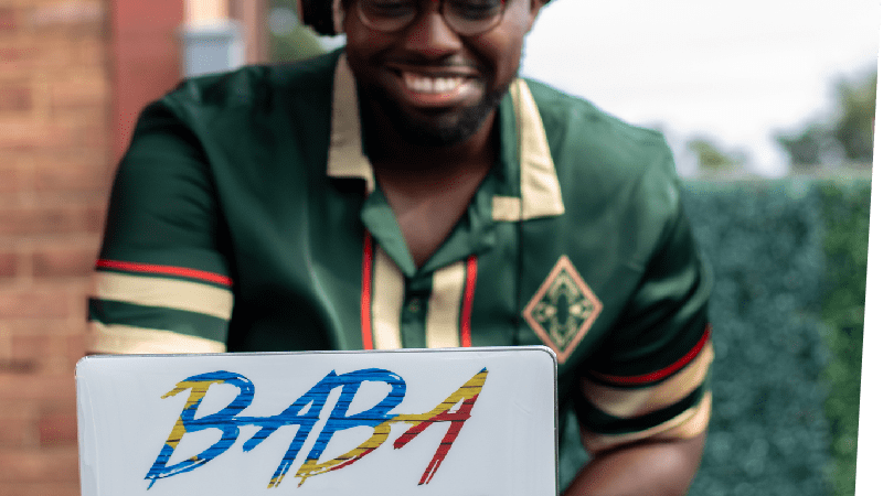 Smiling DJ Baba Yako standing behind a white laptop that displays his name in blue, red, and yellow letters. DJ Baba Yako wears headphones on his head and a green shirt.