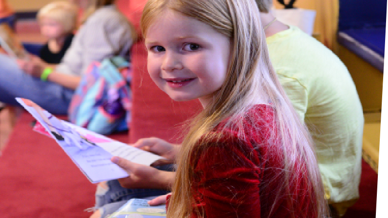Central foreground: Two children attending the LINC storytime reading event. One of the children, a girl, smiles at the camera. Beside her, hidden by her frame, sits a boy reading a book.
