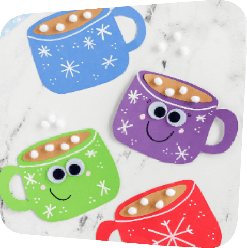 Four hot cocoa magnet crafts on a white countertop. Top left: blue mug with white snowflakes, fake brown "hot chocolate and fluffy white marshmallows" inside. Underneath, from right to left going down: purple, green, and red hot chocolate magnet mugs. The purple and green mugs have googly eyes and smiles.