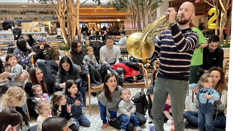 Mozart for Munchkins performer stands before an audience of children and families, playing a horn.