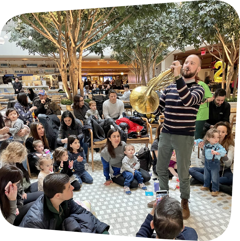Mozart for Munchkins performer stands before an audience of children and families, playing a horn.